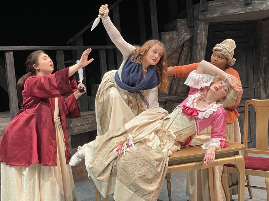 Four actors in period clothing depicting historical women. One is draped across a table, with hand over head in a dramatic gesture, while another woman is climbing the table with a knife held overhead. The two remaining try to hold the woman back.