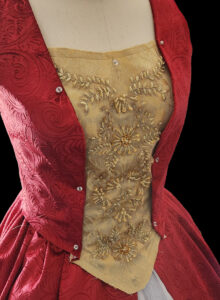 Example of a costume during the building process. The bodice of a dress is being constructed with red fabric on each side and a center panel of gold with detailed embroidery.