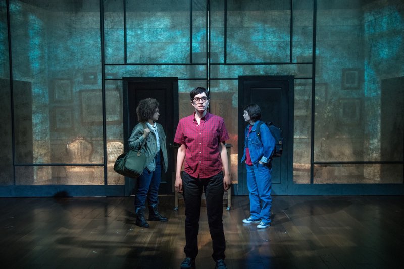 Three actors stand on stage in a triangular formation. One actor is slightly in front of the others and facing forward. The other two actors face each other. The set behind them has two doors and is lit with a teal wash of light.