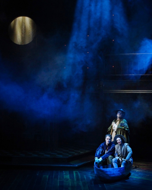 In the bottom left corner of the image, two actors sit in a wooden boat on the floor of a stage with another actor standing directly behind them. Above them, a slight fog is picked up by a blue wash of light. In the top left corner of the image, a moon is seen.