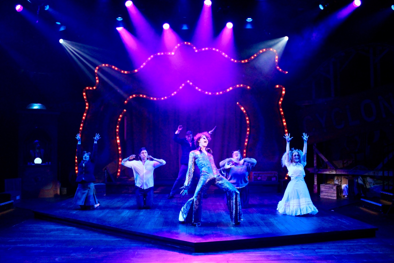 Actors on a stage during a musical number. One actor is in front of the others, who kneel in a line. The central actor wears a black jumpsuit with bellbottoms and sunglasses. He is positioned in a wide stance, leaning slightly back as he sings. Beams of purple light shine down on him.