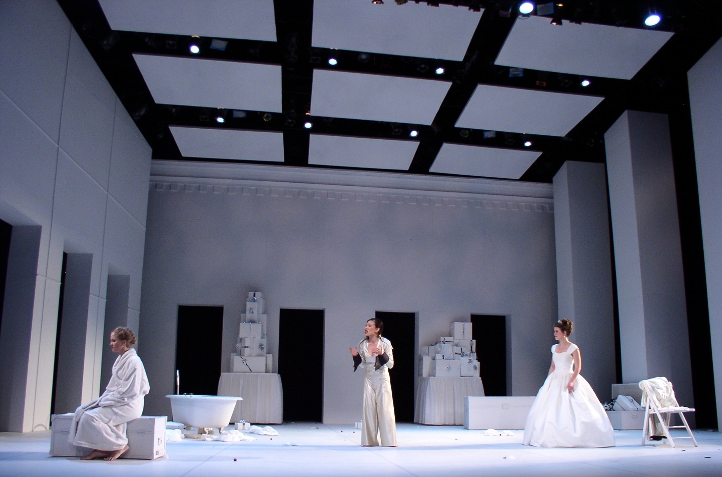 Three actors on a stage made up of white walls. Passageways are evenly spaced along the back wall and the wall on the left side of the stage. Three actors stand equally distant from one another across the stage. They wear white costumes.