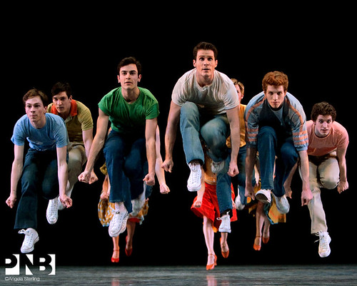 Production photo from "West Side Story." A group of actors are positioned in a small group in the midst of a jump. They all are in the air with their knees up close to their chests, and both arms extended down towards the floor.